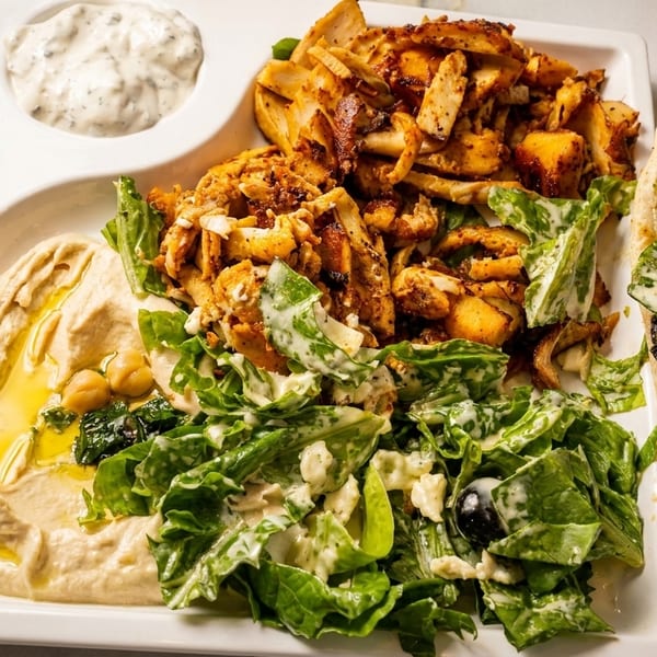 A white platter with chicken shawarma, hummus, salad, tzatziki, and a pita bread sandwich.