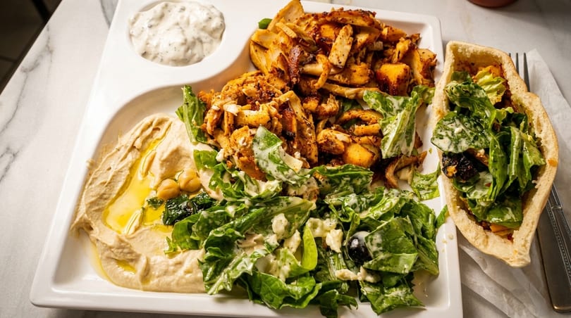 A white platter with chicken shawarma, hummus, salad, tzatziki, and a pita bread sandwich.